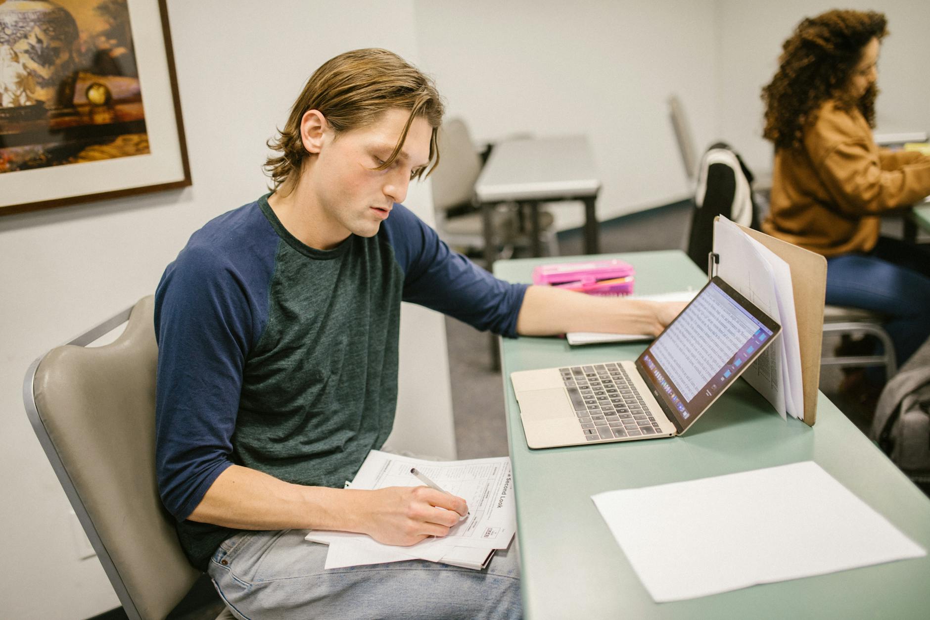 College student writing notes and using a laptop at a classroom desk for exam preparation — TransferCredit.org