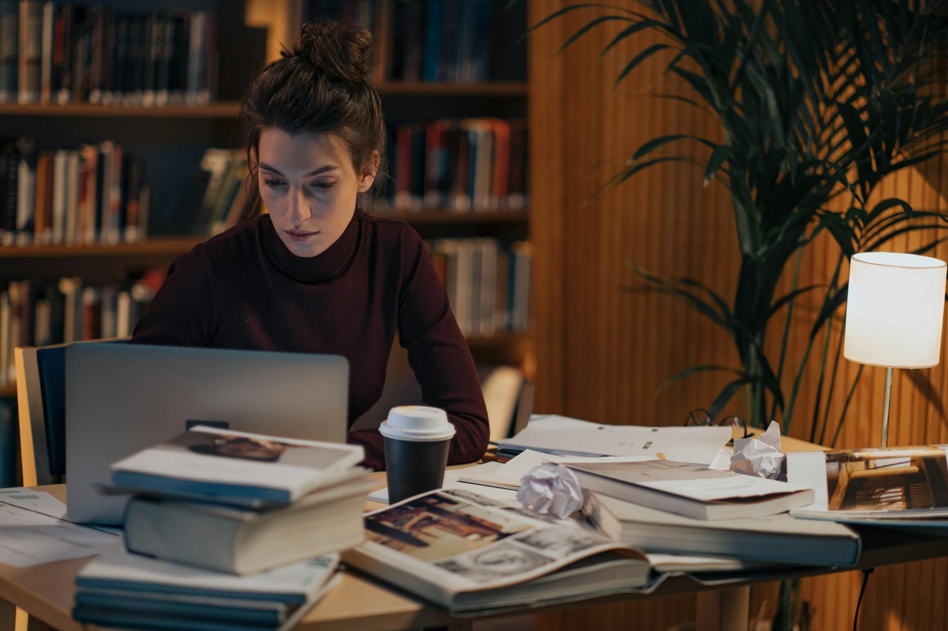 Woman working late at a desk filled with books, laptop, and coffee in a cozy library setting — TransferCredit.org