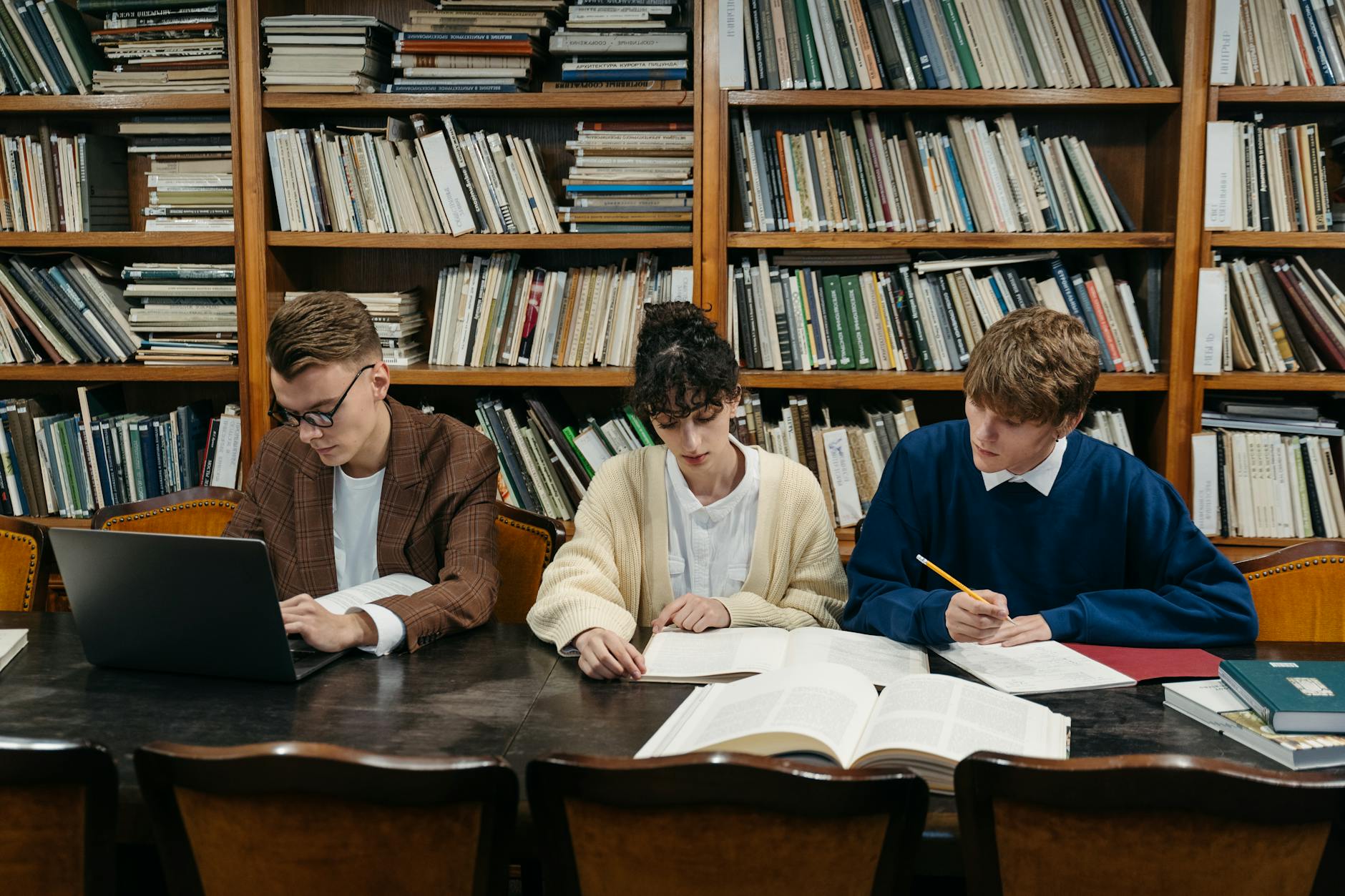 Three students work on laptops and books in a university library filled with shelves — TransferCredit.org