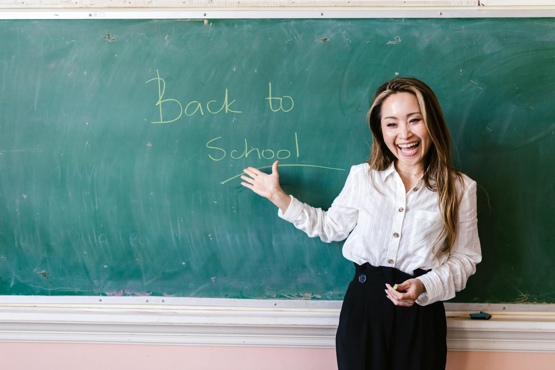 Smiling teacher in a classroom pointing at back to school message on a chalkboard — TransferCredit.org
