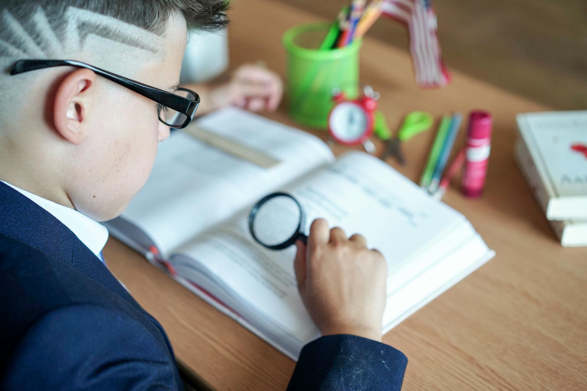 A young boy focuses on his book using a magnifying glass at a study desk — TransferCredit.org