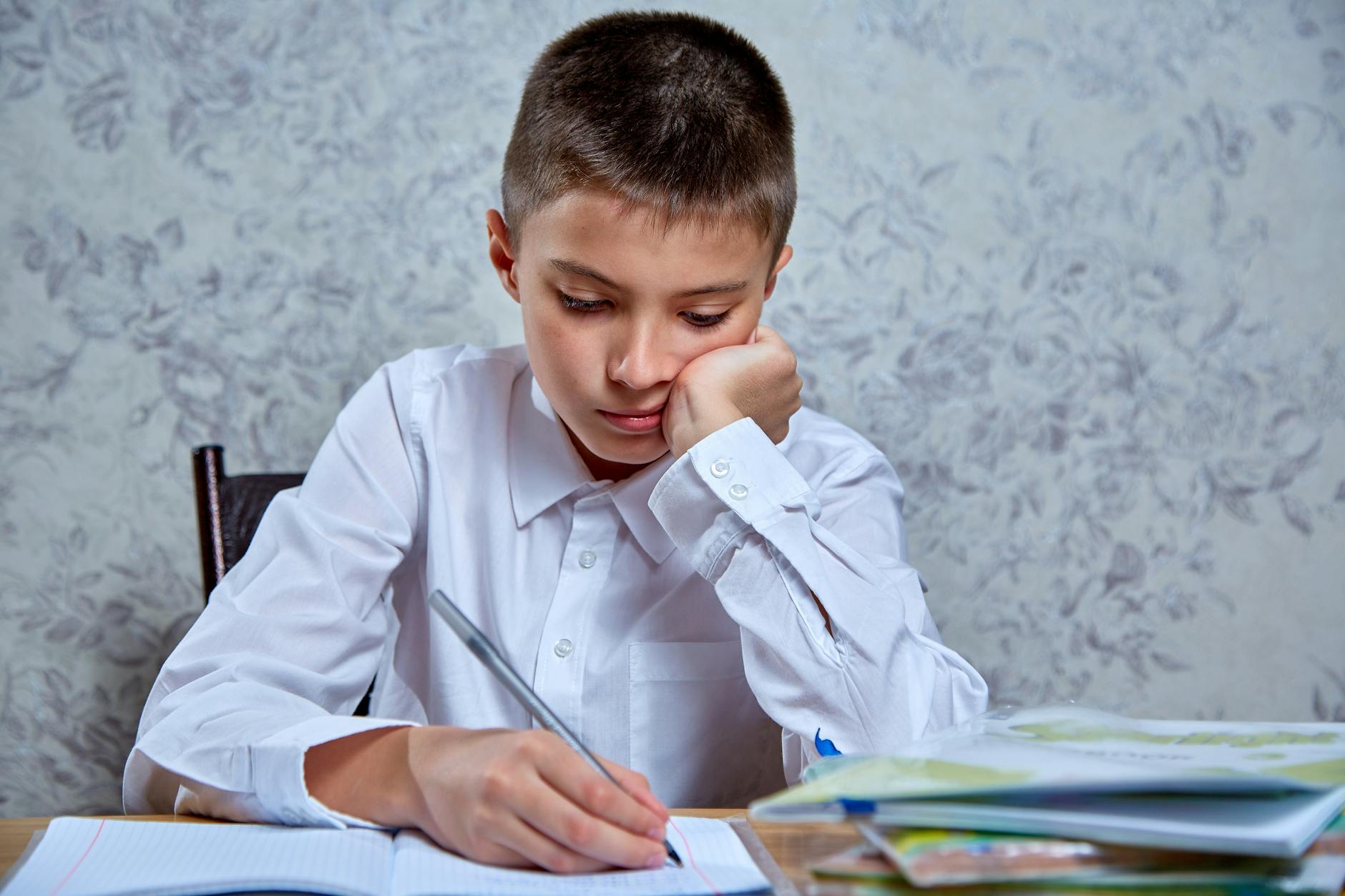 A young boy in a white shirt attentively writing in a notebook at a desk, indoors — TransferCredit.org