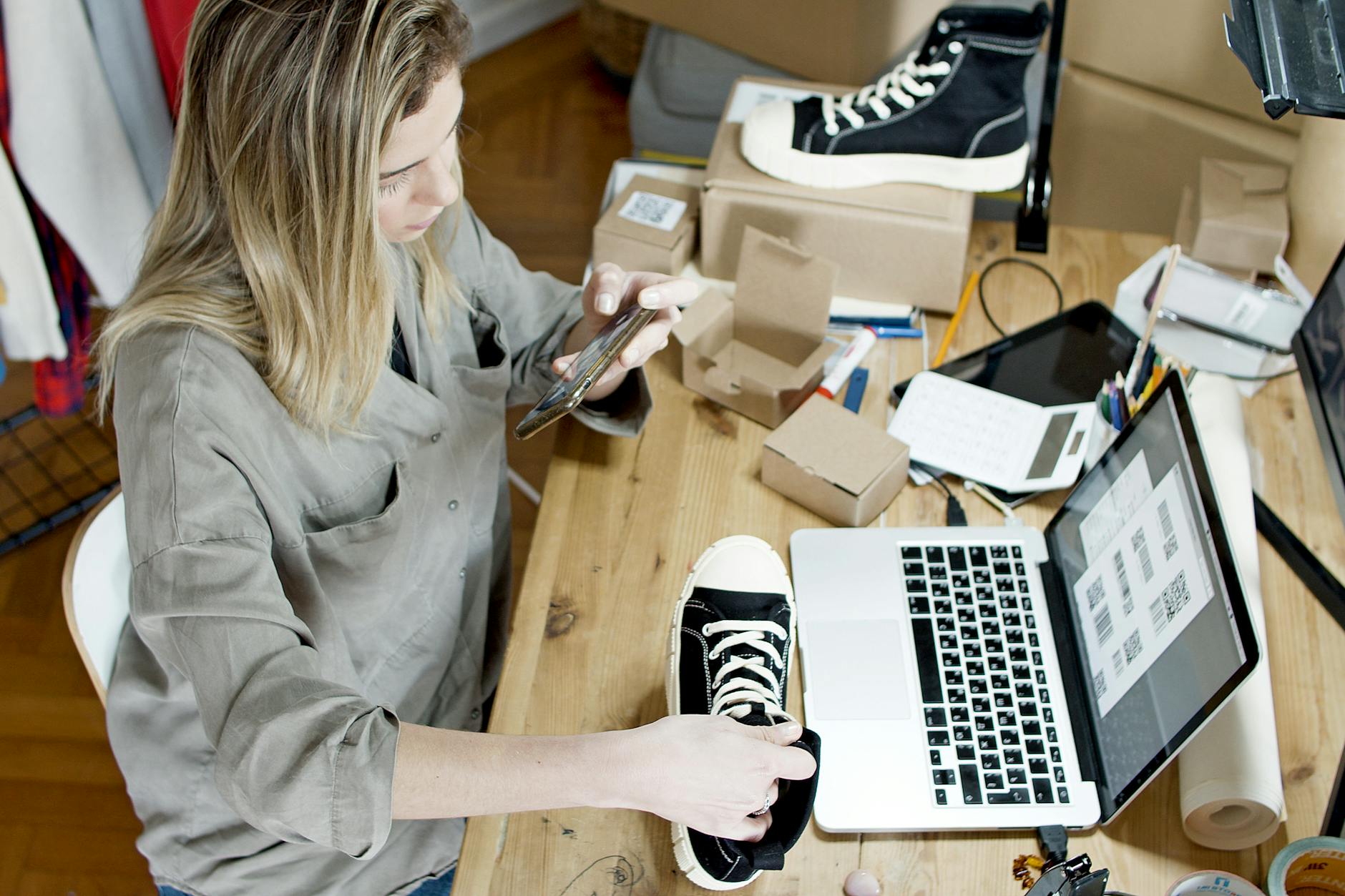 Woman photographing shoes for online sale in home workspace with laptop and packages — TransferCredit.org