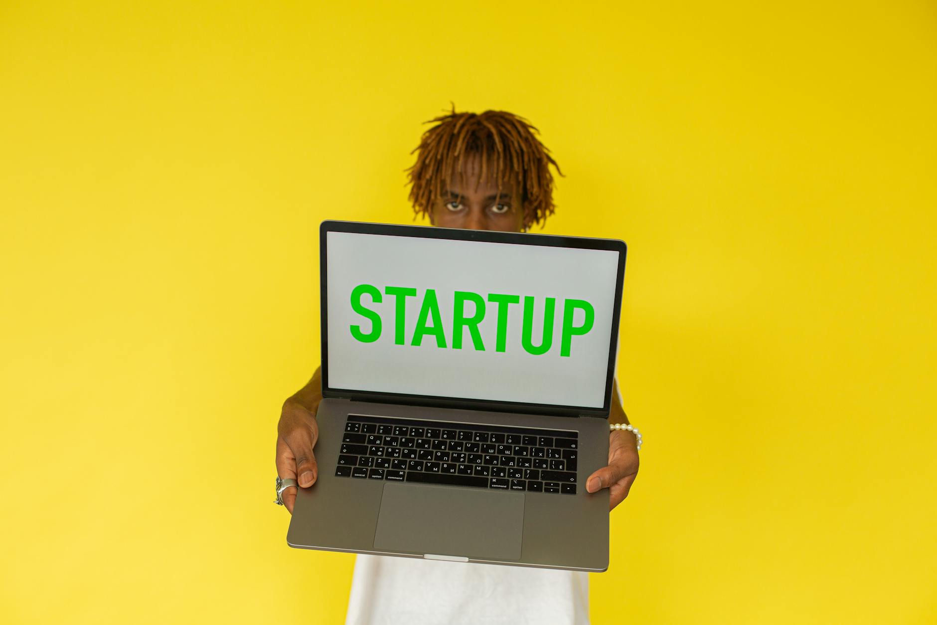 A young black man holds a laptop displaying 'Startup' against a vibrant yellow background — TransferCredit.org