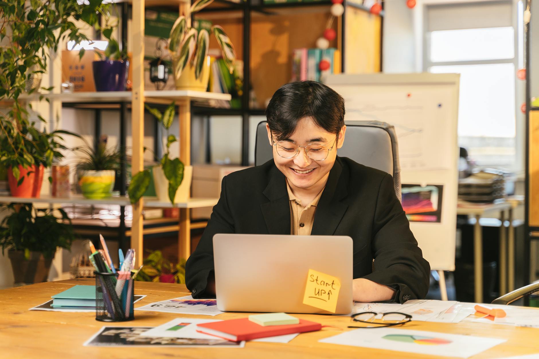 Smiling young professional in black suit working on laptop at a cozy office desk surrounded by plants — TransferCredit.org