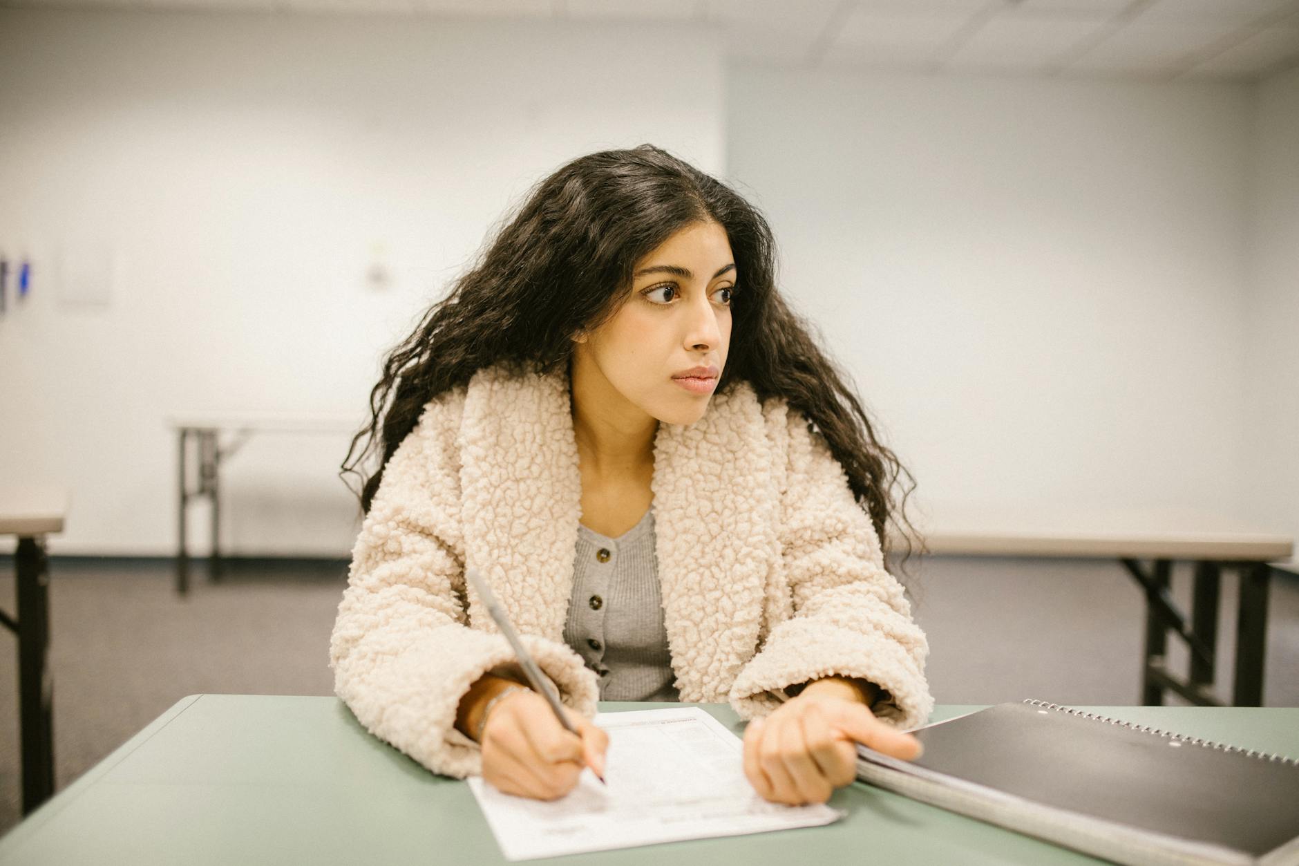 A college student writing on a test paper while looking away in a classroom setting — TransferCredit.org