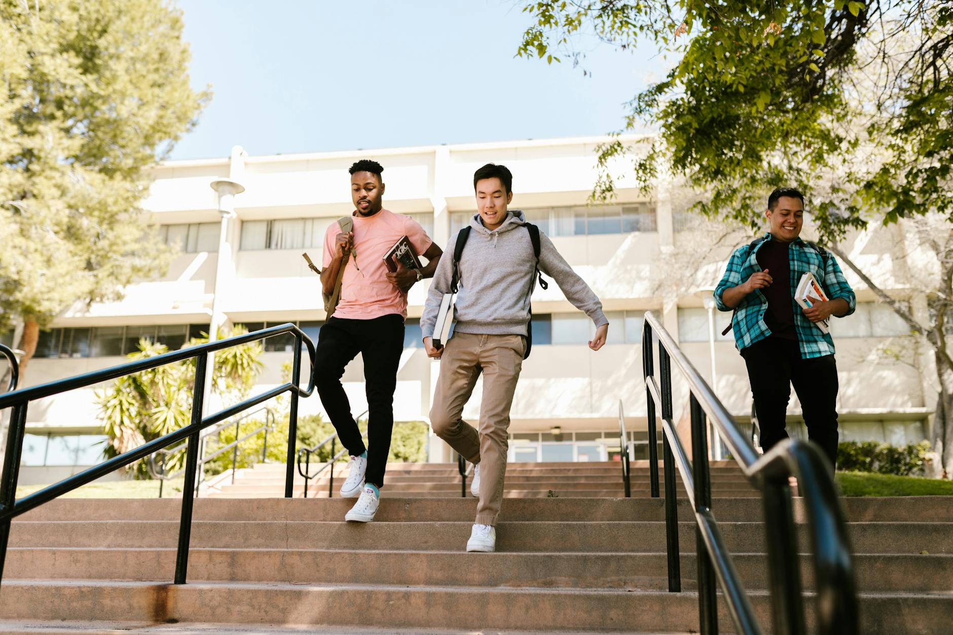 Three college students walk down stairs on campus, carrying books and backpacks, on a sunny day — TransferCredit.org