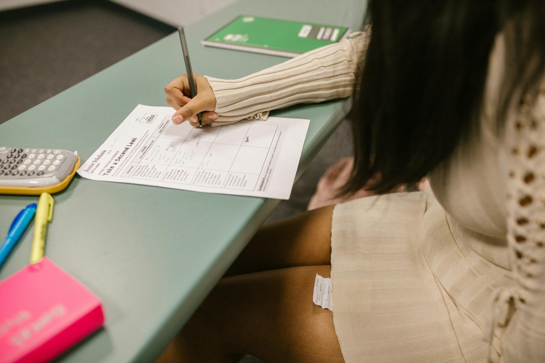 Student secretly looking at a cheat sheet under the desk while taking an exam in a classroom setting — TransferCredit.org