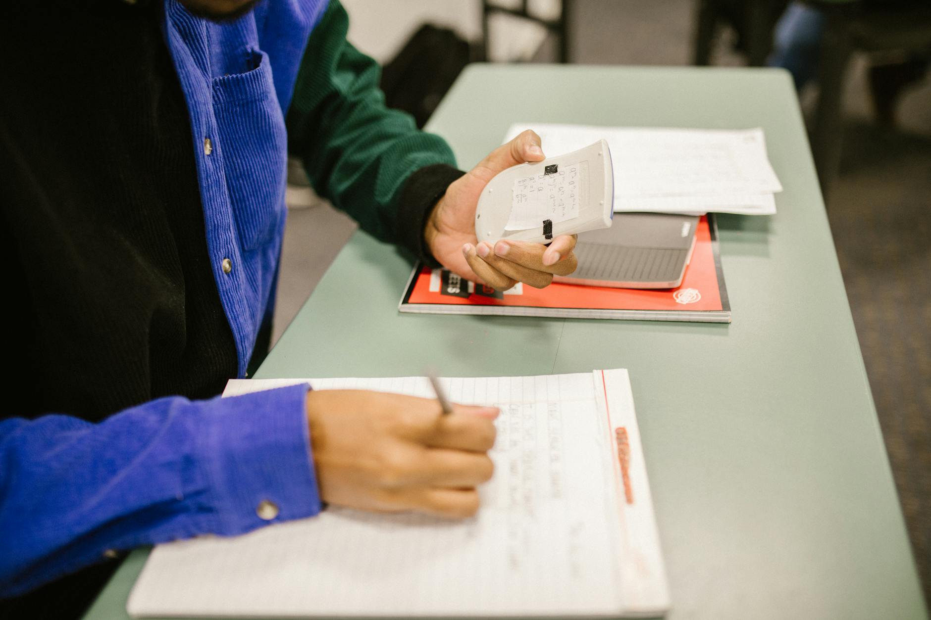 Student using a calculator and taking notes during exam preparation in a classroom — TransferCredit.org