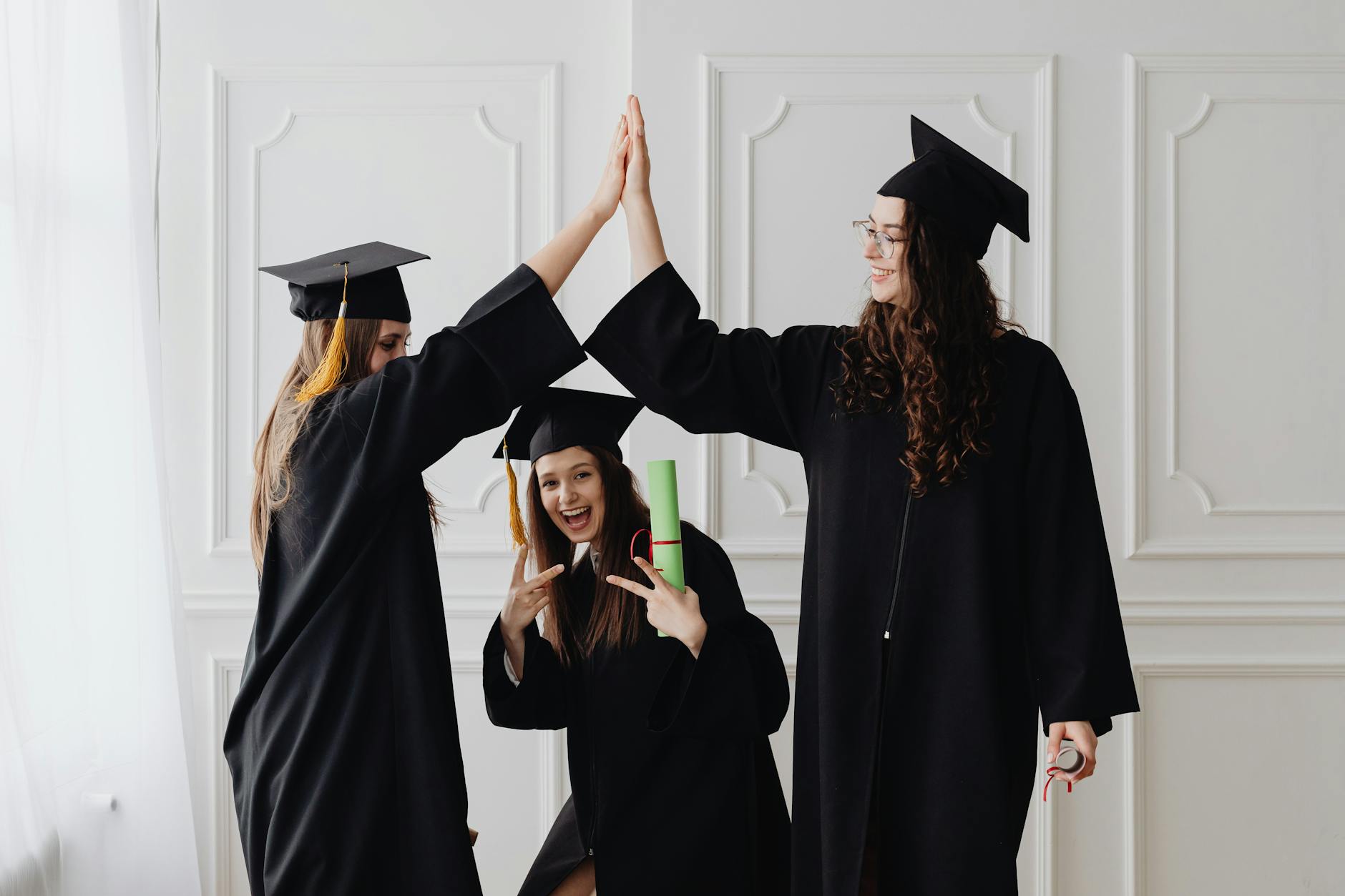Three young women celebrating graduation with a high five, holding diplomas indoors — TransferCredit.org