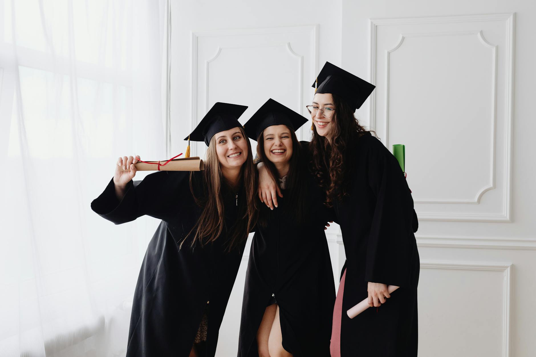 Three smiling graduates in caps and gowns celebrating indoors together — TransferCredit.org
