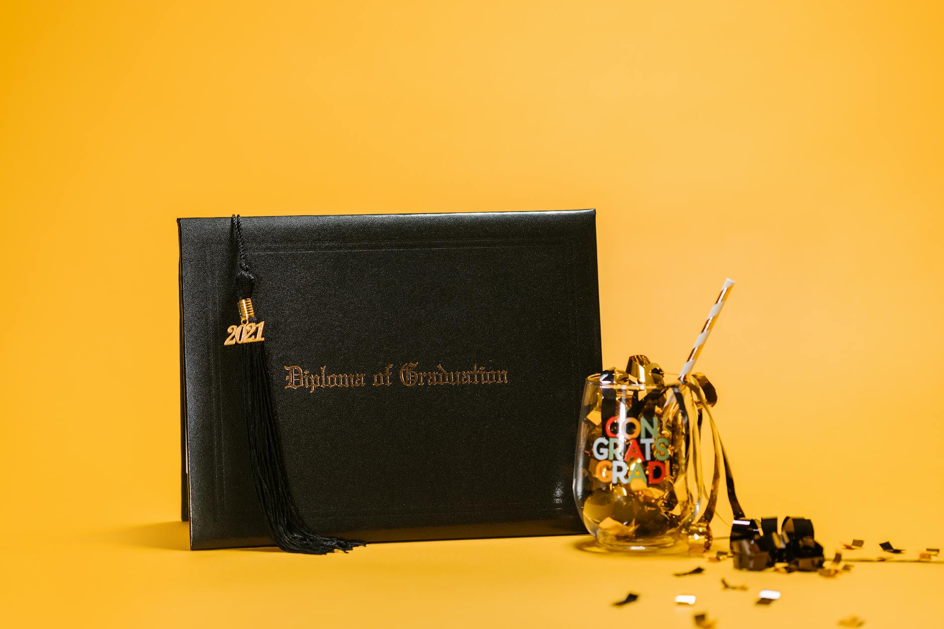 Black diploma and glass with confetti against a yellow background, symbolizing graduation celebration — TransferCredit.org