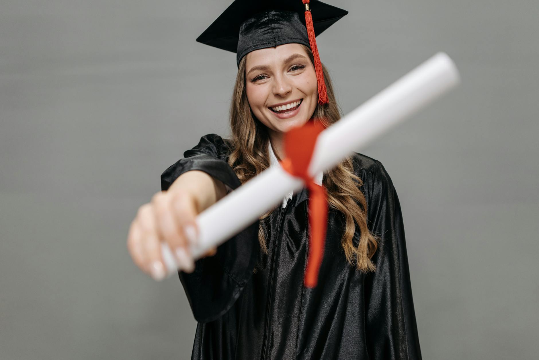 Happy female graduate wearing a cap and gown proudly holding her diploma with a smile — TransferCredit.org