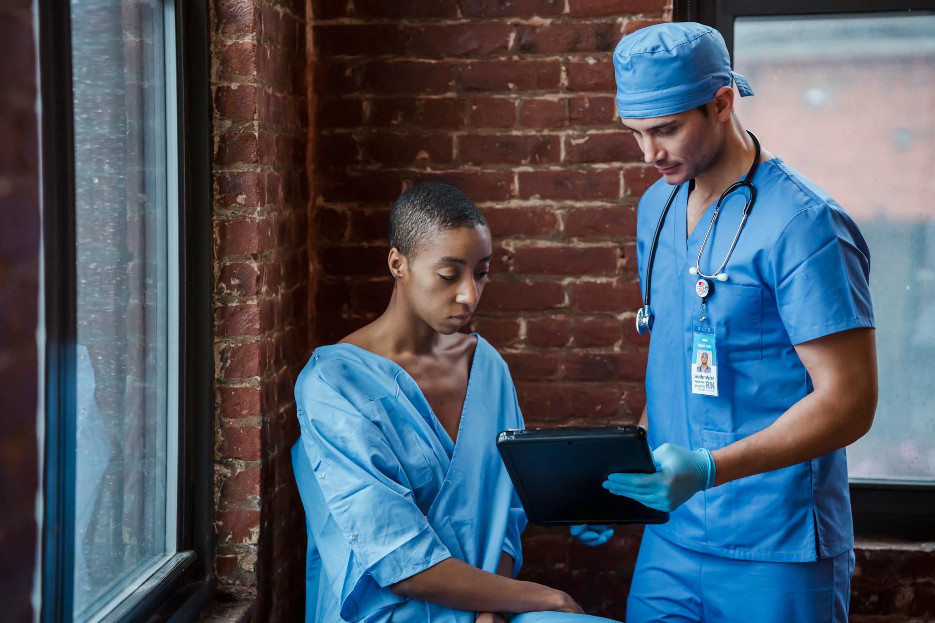 Thoughtful doctor in uniform and gloves showing diagnosis to African American female patient with short hair in blue medical robe in hallway of hospital in daytime — TransferCredit.org