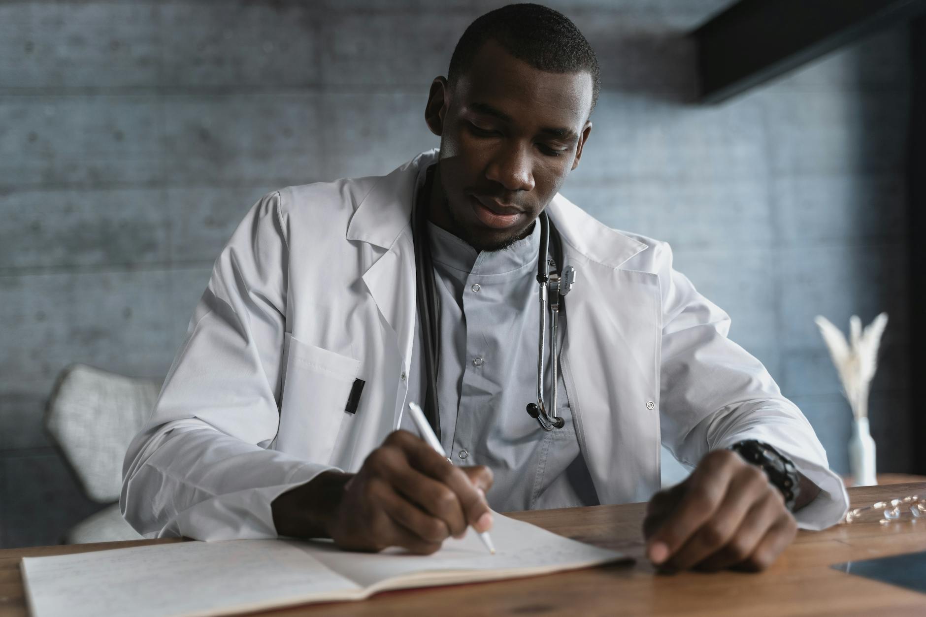 Black male doctor in a lab coat writing notes at a wooden desk in a medical office — TransferCredit.org
