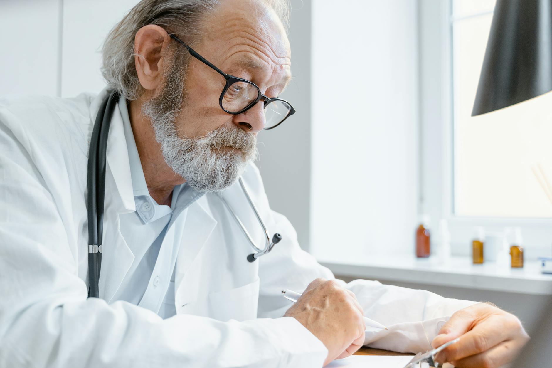 Elderly doctor wearing eyeglasses and stethoscope writes at office desk — TransferCredit.org