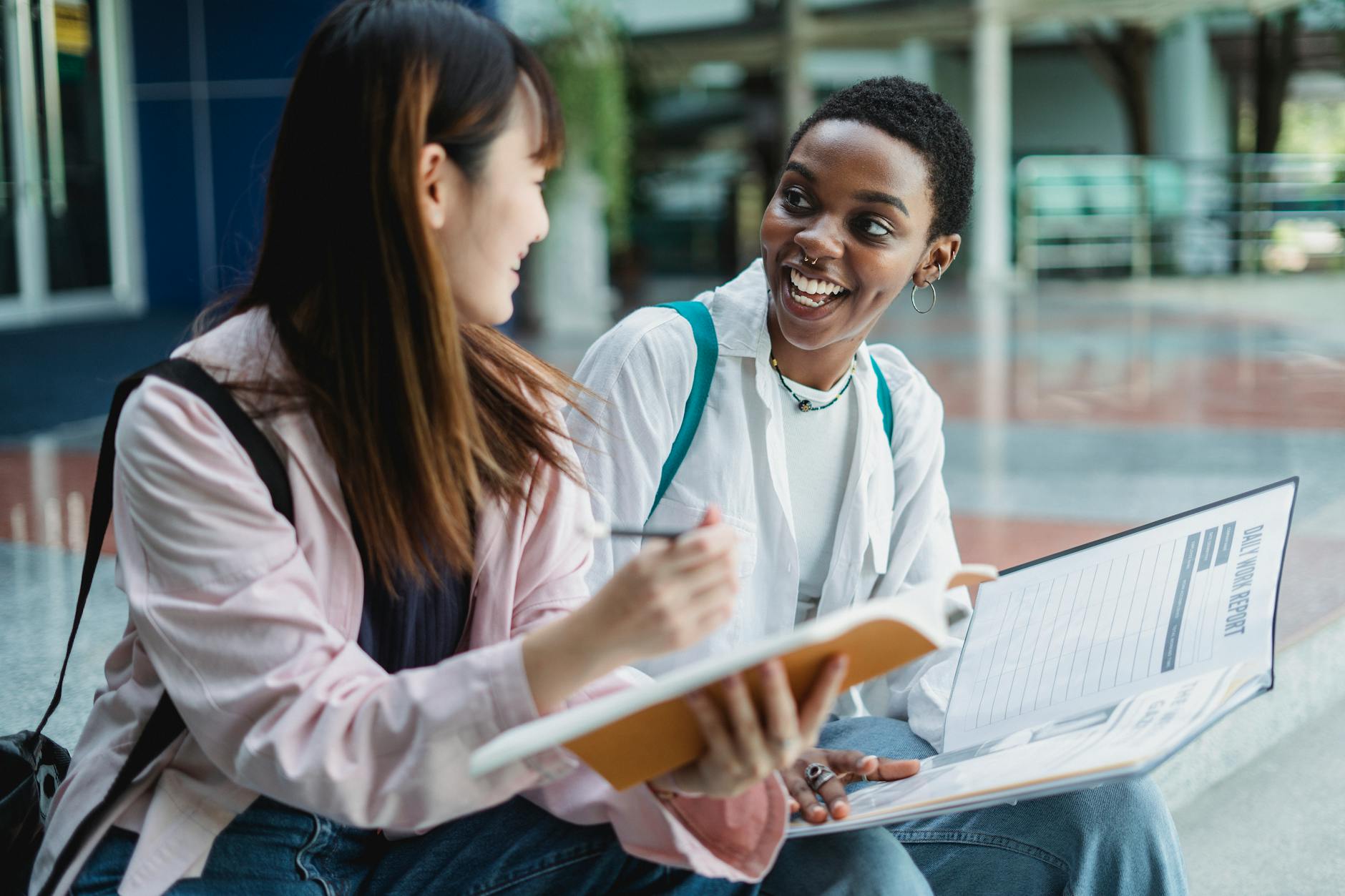 Happy young multiracial women in casual clothes sitting outdoors with notepad and papers in daytime while looking at each other — TransferCredit.org