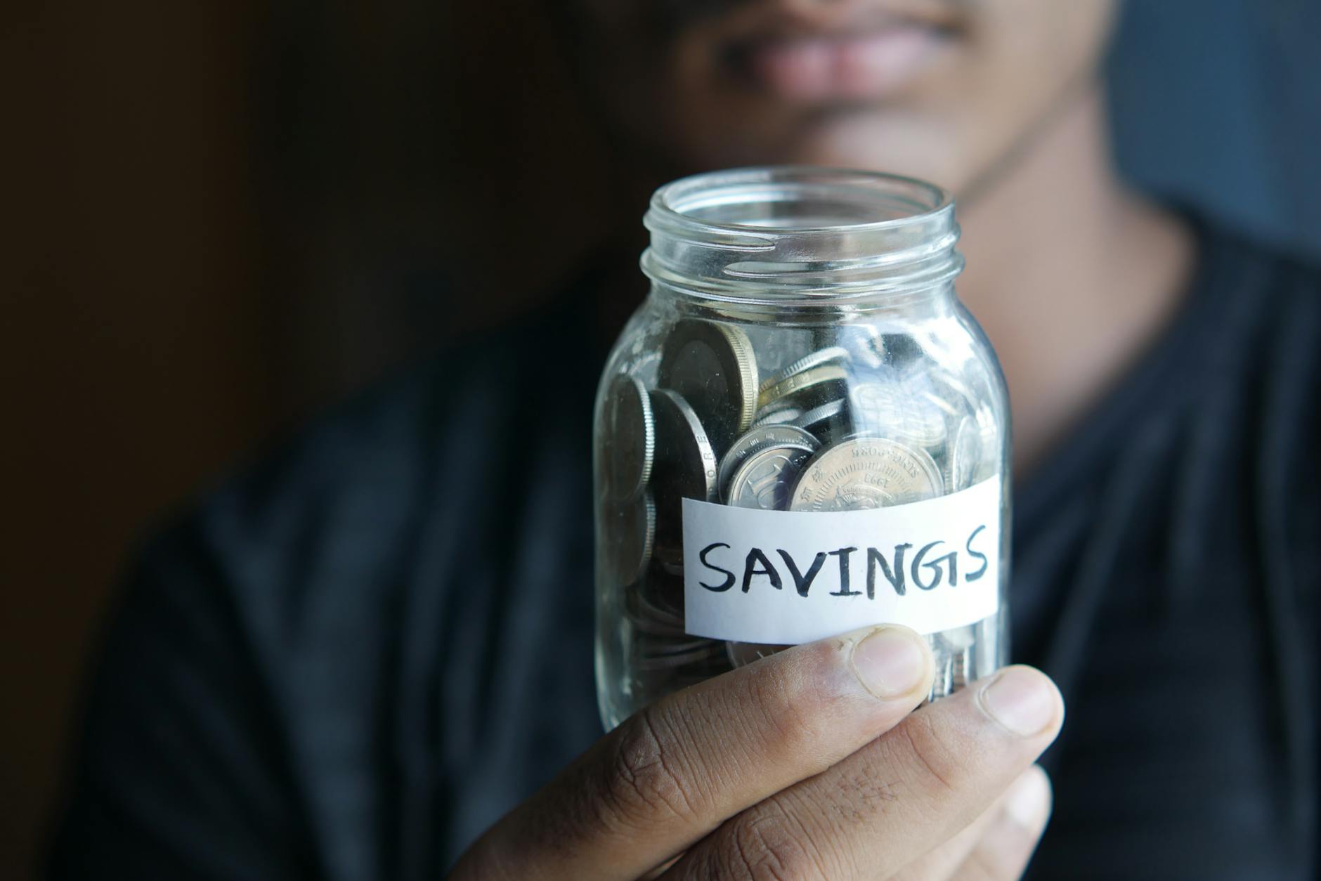 A close-up image of a person's hand holding a jar full of coins labeled 'Savings' — TransferCredit.org