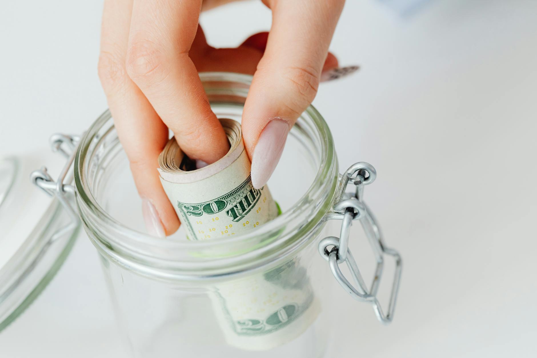 A close-up of a hand placing rolled dollars into a glass jar, symbolizing savings — TransferCredit.org