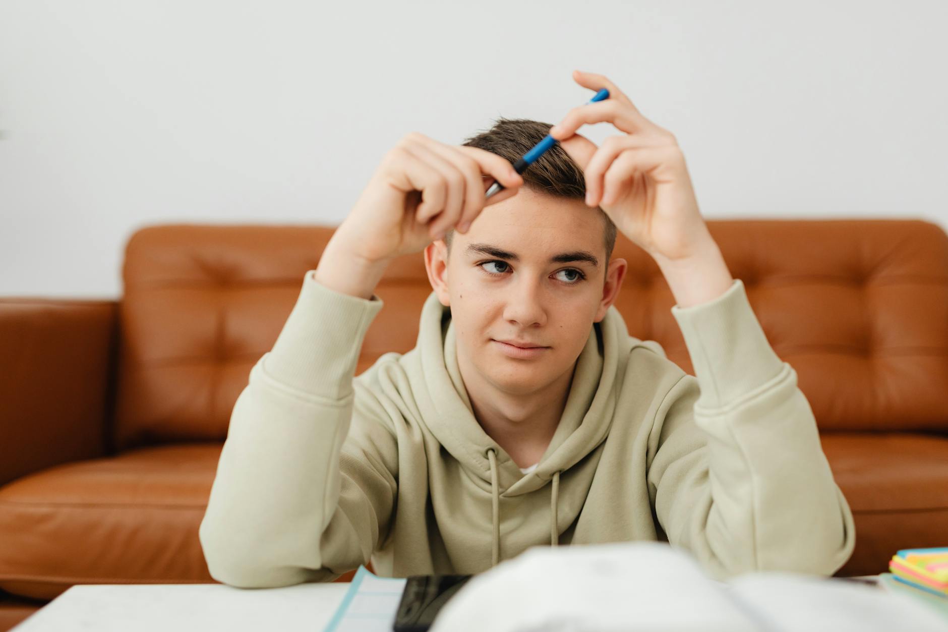 Teenager in hoodie contemplating with pen while studying on a brown sofa indoors — TransferCredit.org