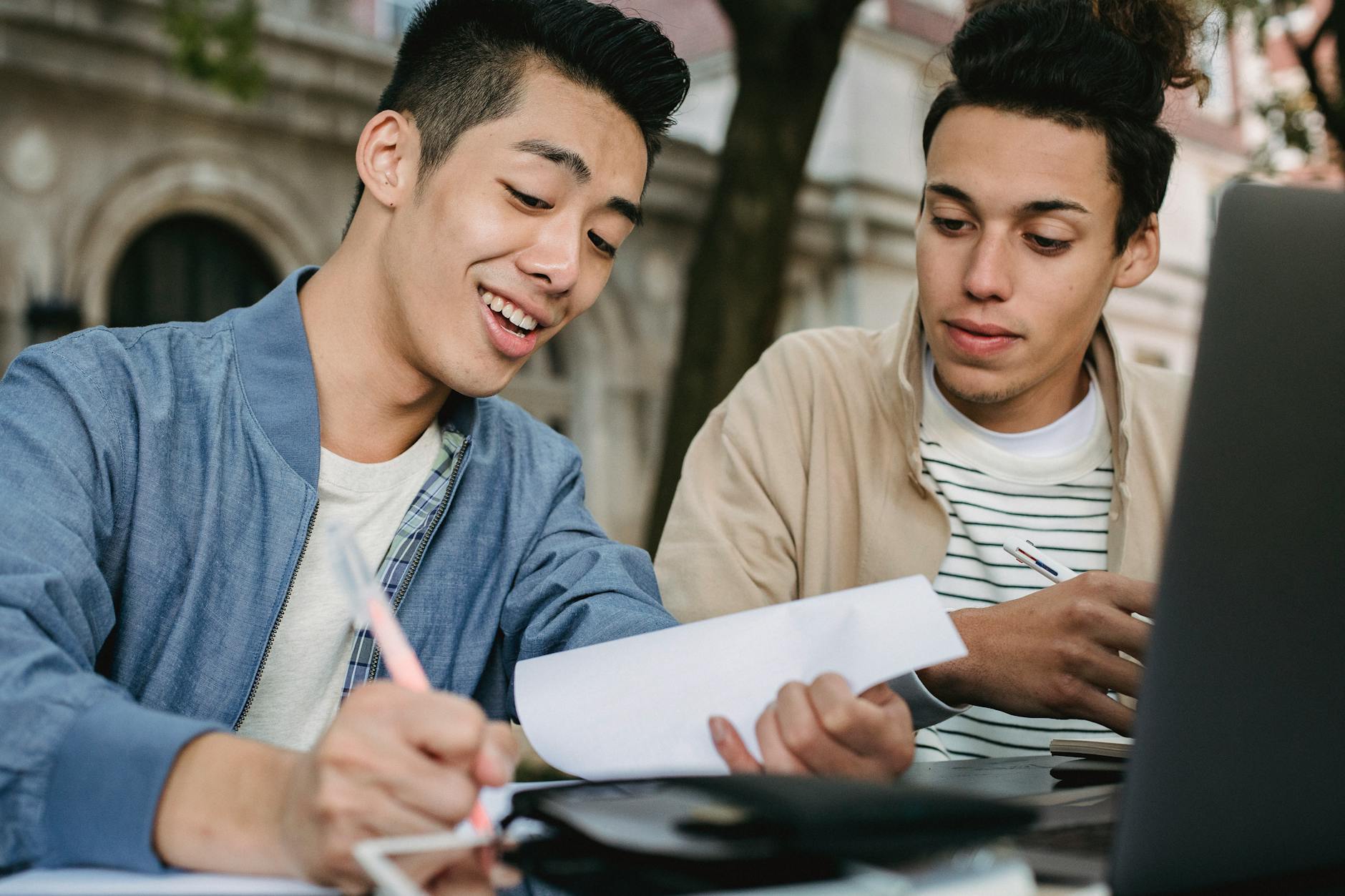 Smiling ethnic male learner writing in document while doing homework assignment with classmate in park — TransferCredit.org