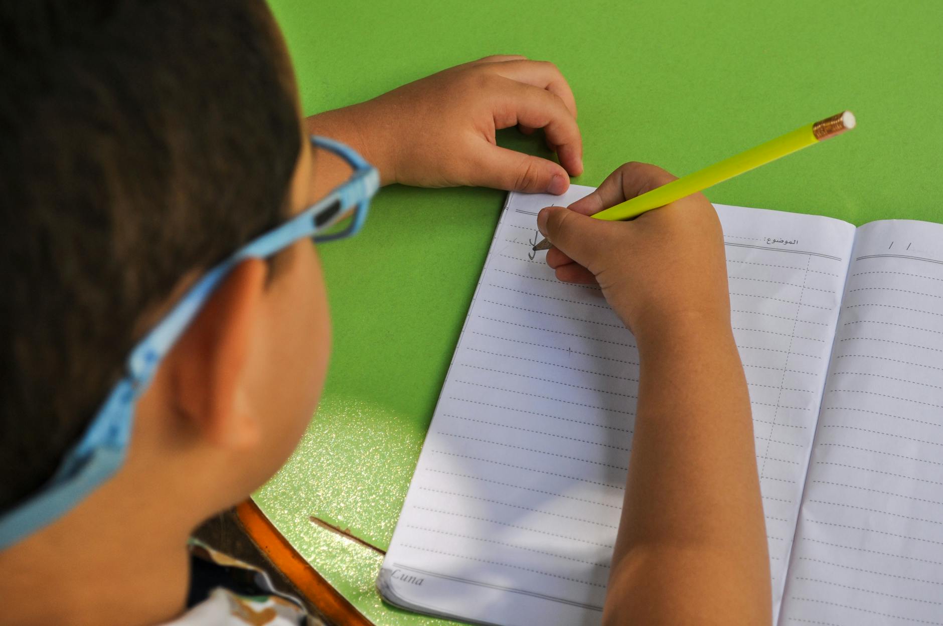 A child wearing glasses writes in a lined notebook with a yellow pencil on a green table — TransferCredit.org