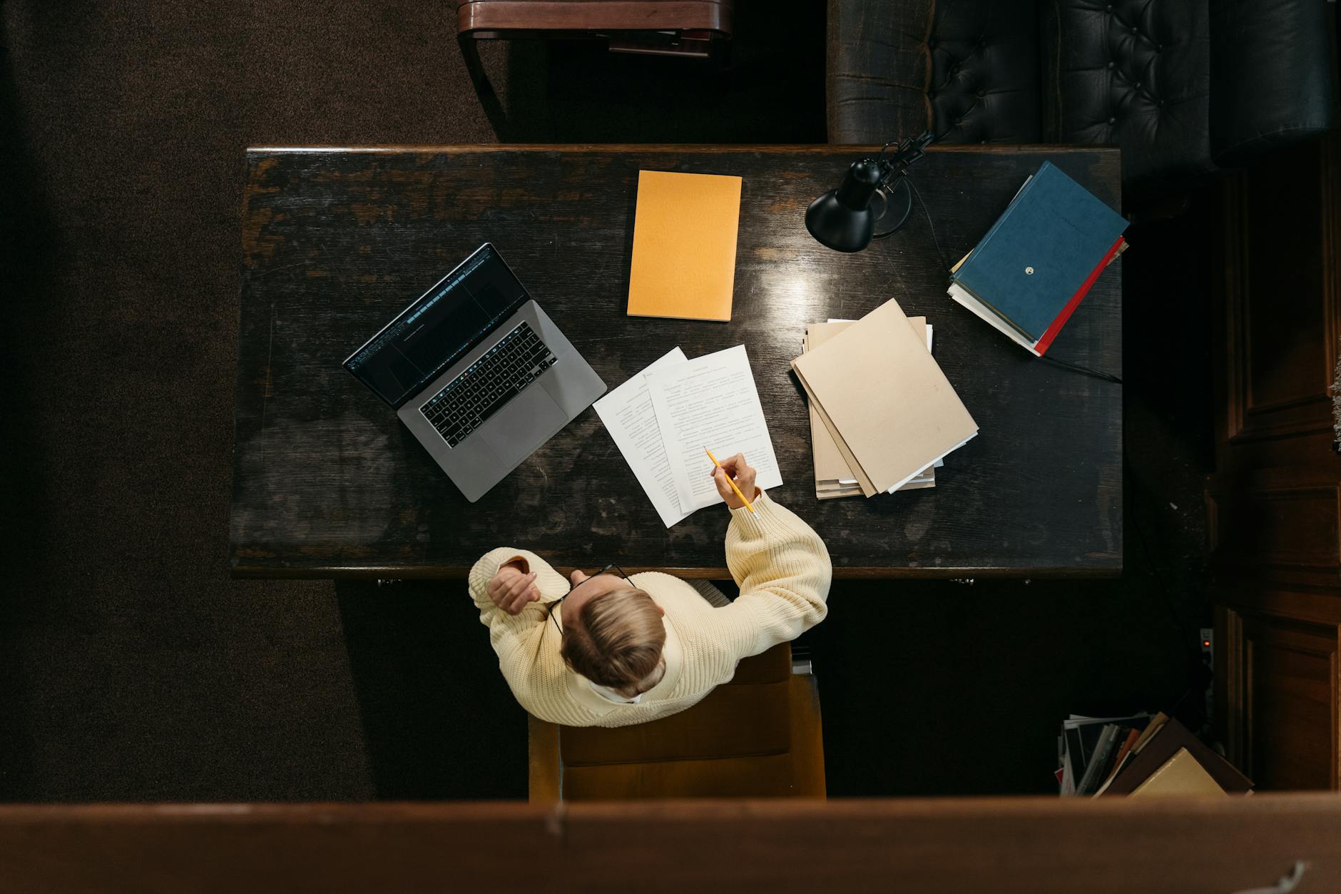 Person in yellow sweater working at a wooden desk with documents, folders, and a laptop — TransferCredit.org